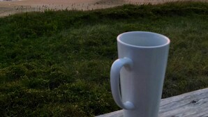 Een witte beker op een houten leuning met uitzicht op een strand bij zonsondergang.