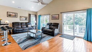 A spacious living room with a black leather sofa, a glass coffee table, and a ceiling fan. There are two large windows with blue curtains, and a balcony is visible through the windows. - Beachside Home • Fenced Yard • Dogs Welcome (Norfolk)