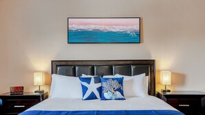 A neatly made bed with white and blue bedding, two decorative pillows, a digital clock, bedside lamps, and a framed picture of a beach scene on the wall. - Beachside Home • Fenced Yard • Dogs Welcome (Norfolk)