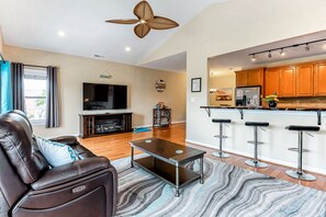 A modern living room with a leather sofa, a coffee table, and a TV. The kitchen has wooden cabinets and a bar area with stools.