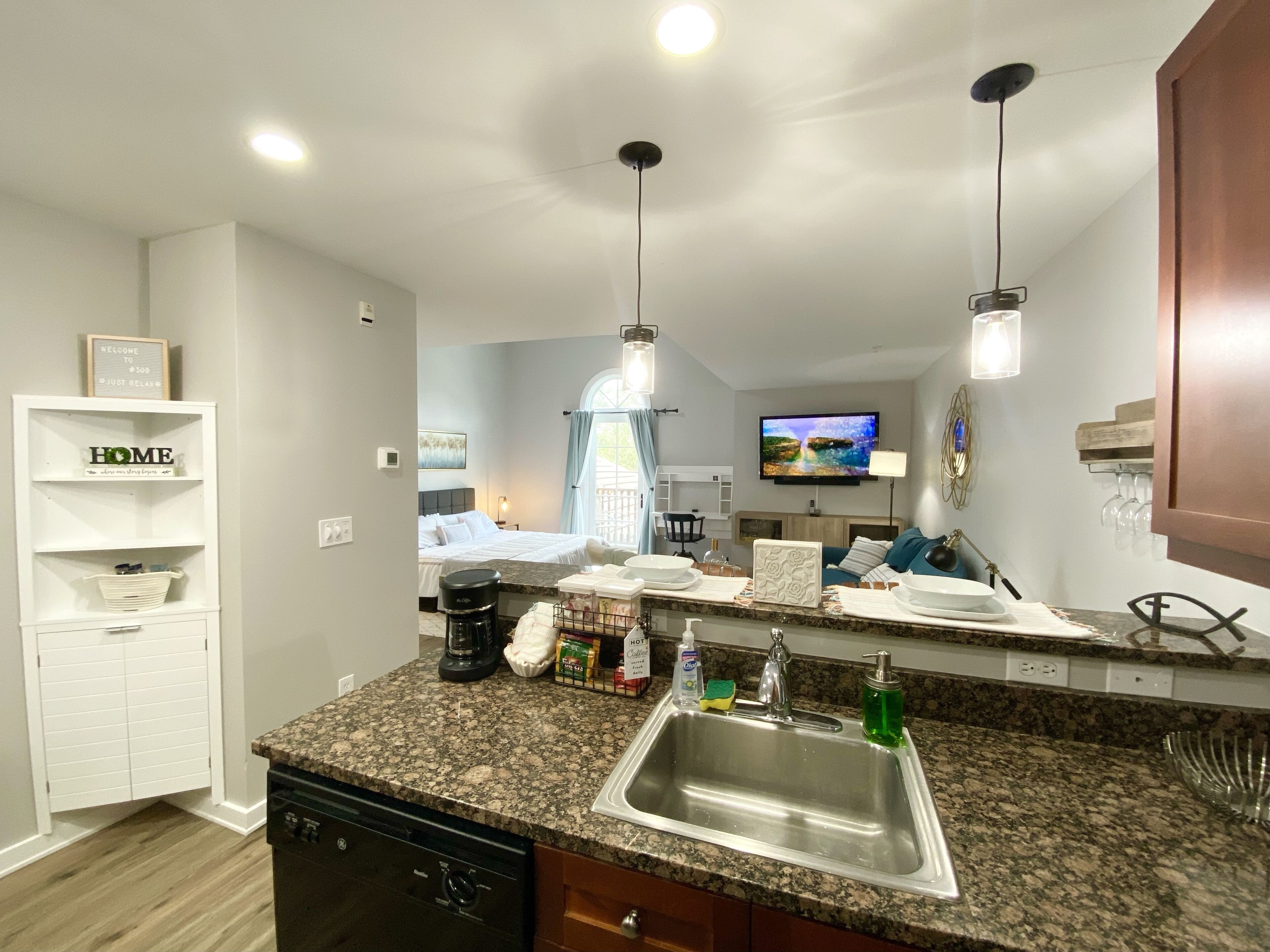 A modern kitchen with granite countertops, a stainless steel sink, and a dishwasher. There is a dining area with a table and chairs, and a TV mounted on the wall.