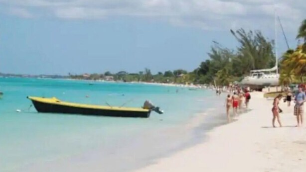 A beach with a yellow boat, clear turquoise water, and people walking on the sand.