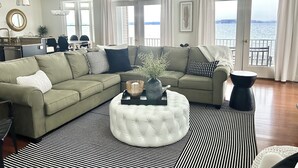 A living room with a sectional sofa, a round ottoman, and a view of the water through large windows.