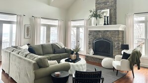 A spacious living room with a high ceiling, a fireplace, a large sofa, and a view of the outdoors.