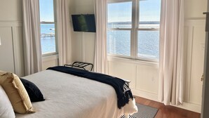 A bedroom with a bed, a TV mounted on the wall, and a view of the sea through large windows.