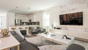 A modern living room with a sofa, coffee table, and TV. A kitchen is visible in the background. - 10BR Villa Near Disney, Pool and Resort Waterpark (Kissimmee)