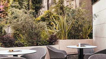An outdoor patio area with round tables and chairs, surrounded by lush greenery and a building with classical architecture.