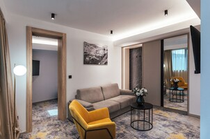 Quarto de hotel moderno com sofá, poltrona amarela, mesa pequena com um vaso, quadro na parede e um espelho grande que reflete o quarto.
