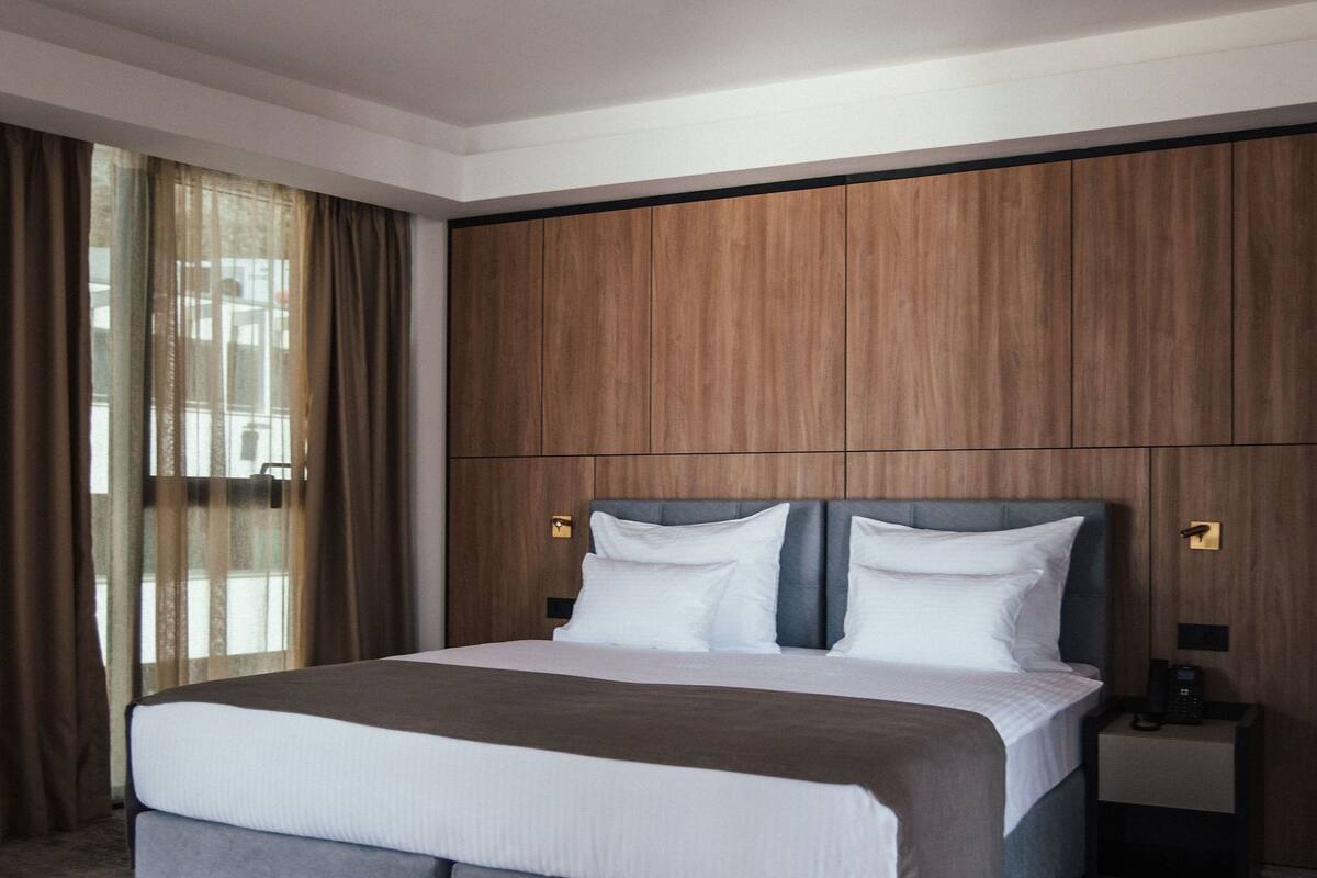 Ein modernes Hotelzimmer mit einem großen Bett, Holzvertäfelung an den Wänden, einem runden Beistelltisch und Blick durch ein Fenster mit Vorhängen.