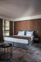 Quarto de hotel moderno com uma cama grande, painéis de madeira nas paredes, uma mesa lateral redonda e vista pela janela com cortinas.