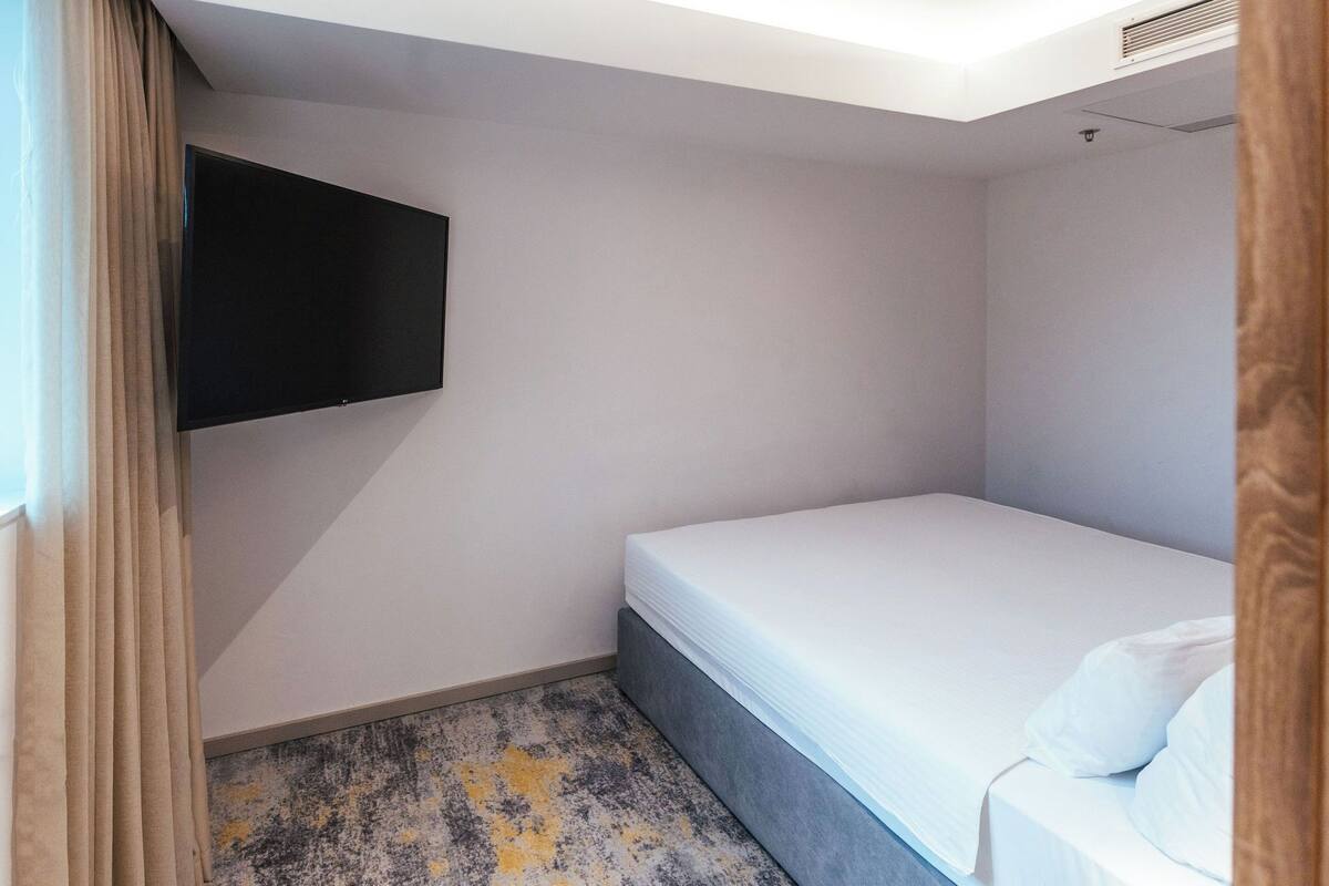Ein kleines Zimmer mit einem Einzelbett, einem flachbildfernseher an der Wand, einem Fenster mit Vorhängen und einem gemusterten Teppich.