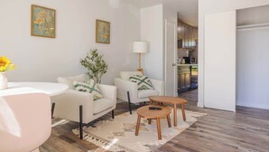 Un salon moderne avec une table ronde, deux canapés, une table basse et une cuisine visible à travers une porte ouverte.