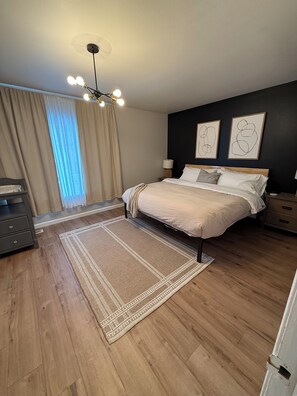 Une chambre Ă coucher comprenant un lit, des tables de chevet, une commode, une fenĂȘtre avec des rideaux et un luminaire au plafond.