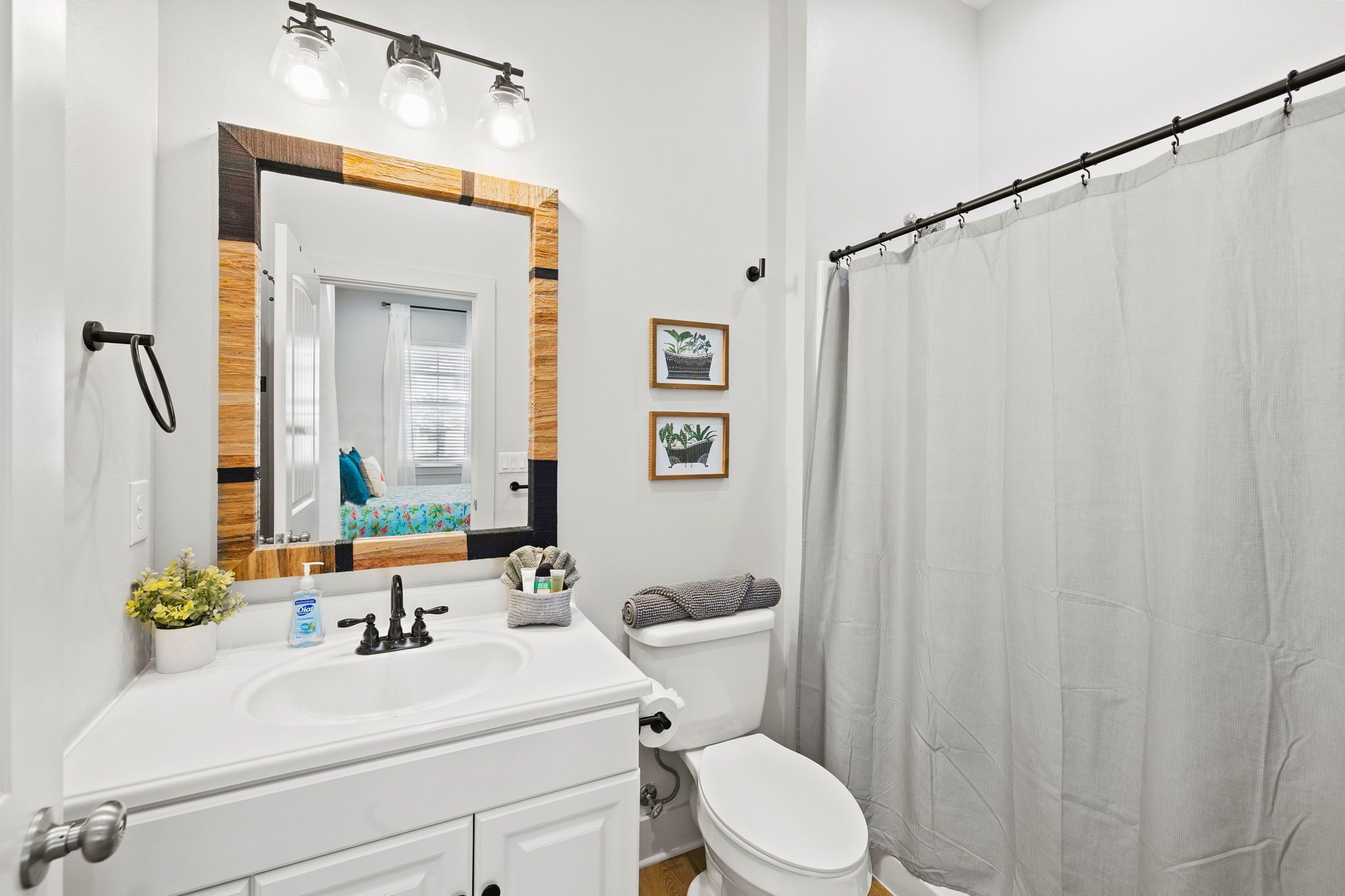 Un bagno con lavandino, specchio e water bianchi. Due quadri appesi alla parete e una tenda da doccia.