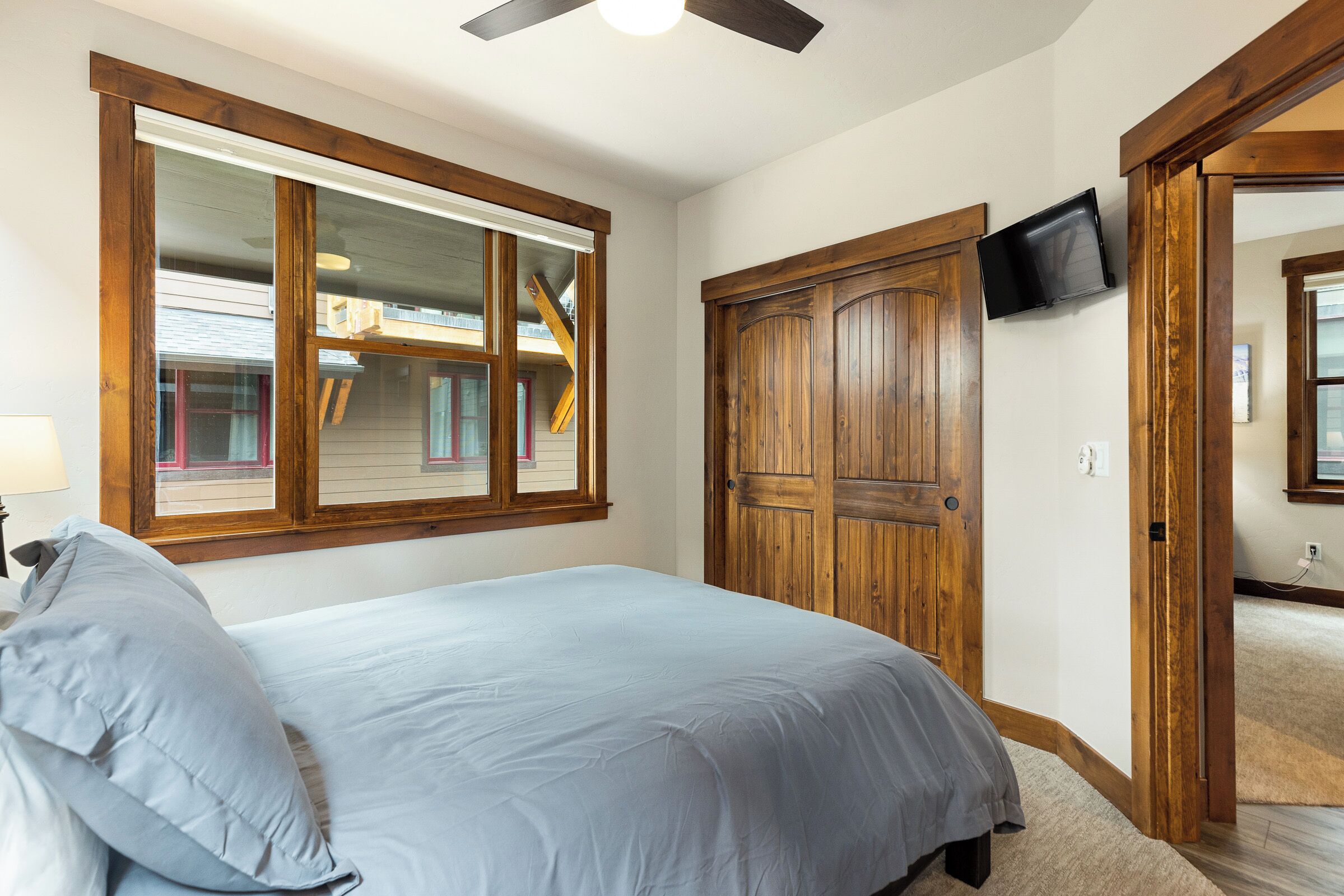 A bedroom with a bed, a wooden door, a TV mounted on the wall, and a window with a view of a house.
