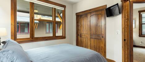 A bedroom with a bed, a wooden door, a TV mounted on the wall, and a window with a view of a house.