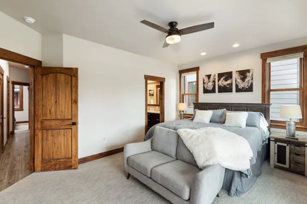 A bedroom with a bed, a gray sofa, a wooden door, and a ceiling fan.