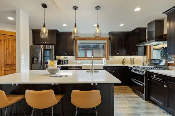 Une cuisine moderne avec des meubles de couleur sombre, un grand îlot central doté de tabourets, un réfrigérateur, une cuisinière et des luminaires suspendus.