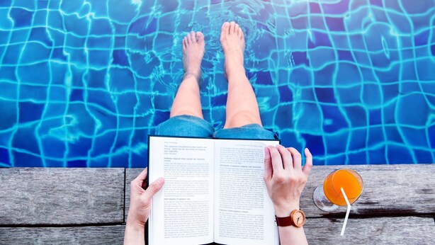 Una persona relaxant-se al costat d'una piscina amb un llibre i una beguda.