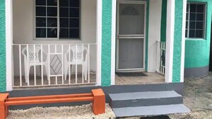 Un edificio con facciata verde e rifiniture bianche. Sono presenti due finestre con cornici bianche, una con vetro a griglia e l'altra con un'unica lastra. Un parapetto bianco circonda una zona relax con due sedie.