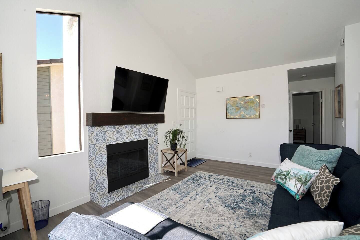 A living room with a fireplace, a flat-screen TV mounted above it, a dark-colored sofa with patterned cushions, and a large window providing natural light.