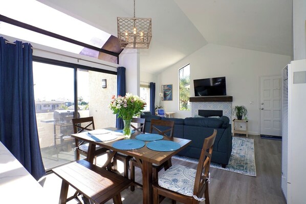 A modern living room with a dining area, a TV mounted on the wall, and a large window with curtains.