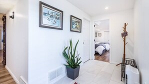 Un couloir avec une plante en pot, des tableaux encadrés, un porte-manteau et, au fond, on aperçoit une chambre avec un lit.