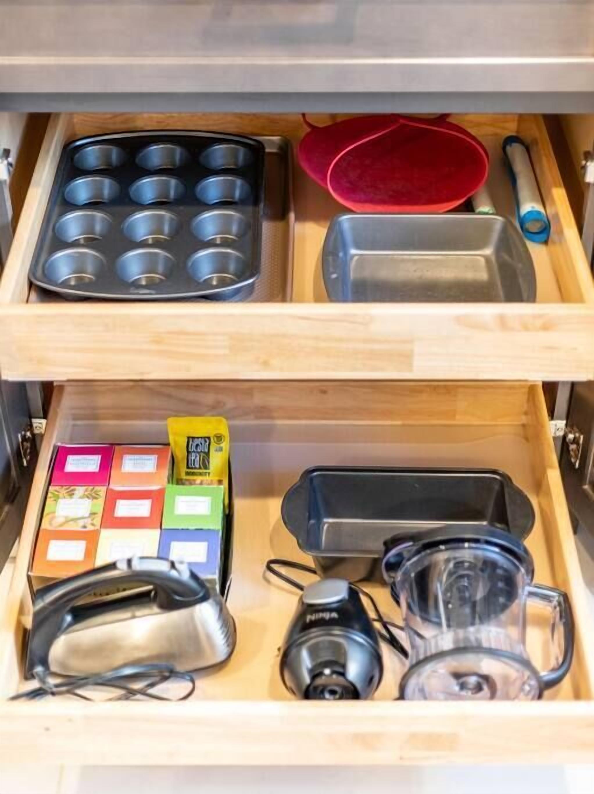 Un cajón de cocina con una bandeja para muffins, una espátula roja, un recipiente de vidrio, una cafetera y varias cajas de té.