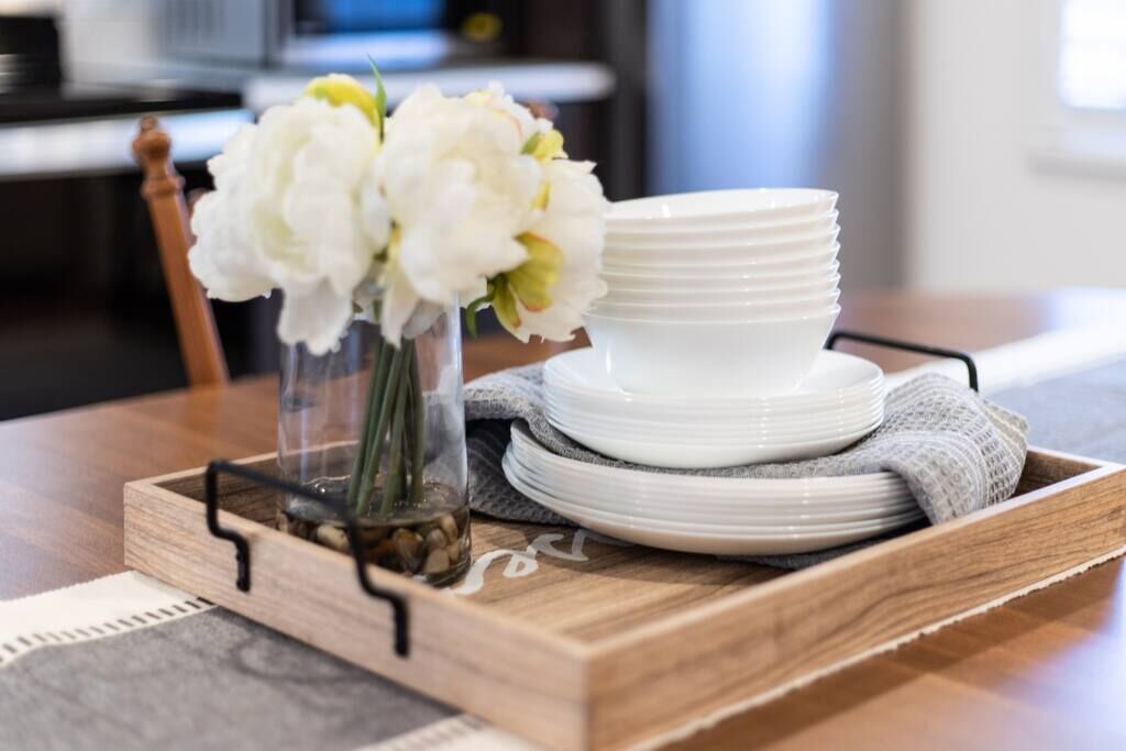 Un plateau en bois sur lequel sont empilées des assiettes blanches, et un vase rempli de fleurs blanches, le tout posé sur une table.