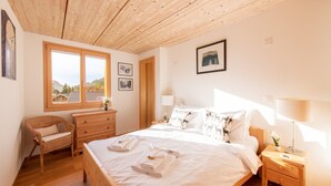 A bedroom with a wooden ceiling, a large bed with white linens, a wicker chair, a wooden dresser, and a nightstand with a lamp.