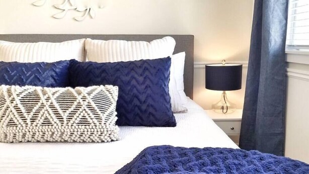 Una cama bien hecha con una colcha blanca y azul estampada, un plaid azul tejido y almohadones decorativos.