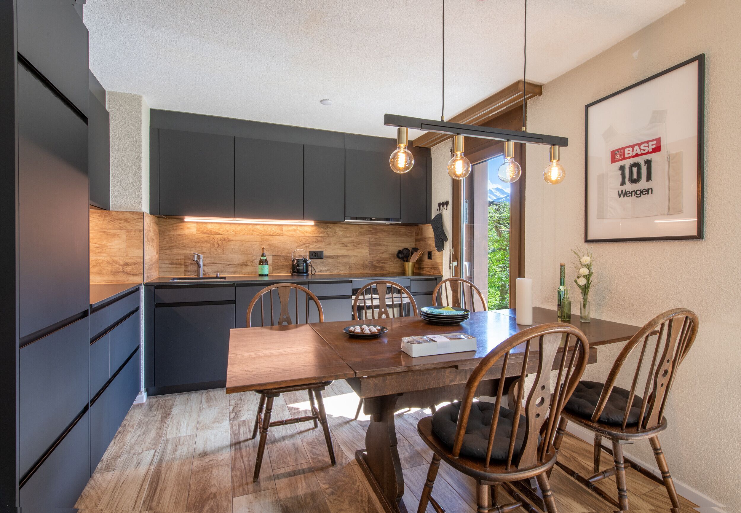 Une cuisine moderne avec des meubles de couleur sombre, une table à manger en bois et un luminaire suspendu.