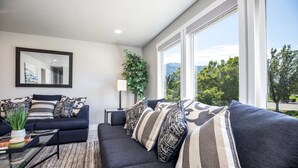 A modern living room with a dark sofa, striped cushions, a glass coffee table, and a large window offering a view of greenery and mountains.