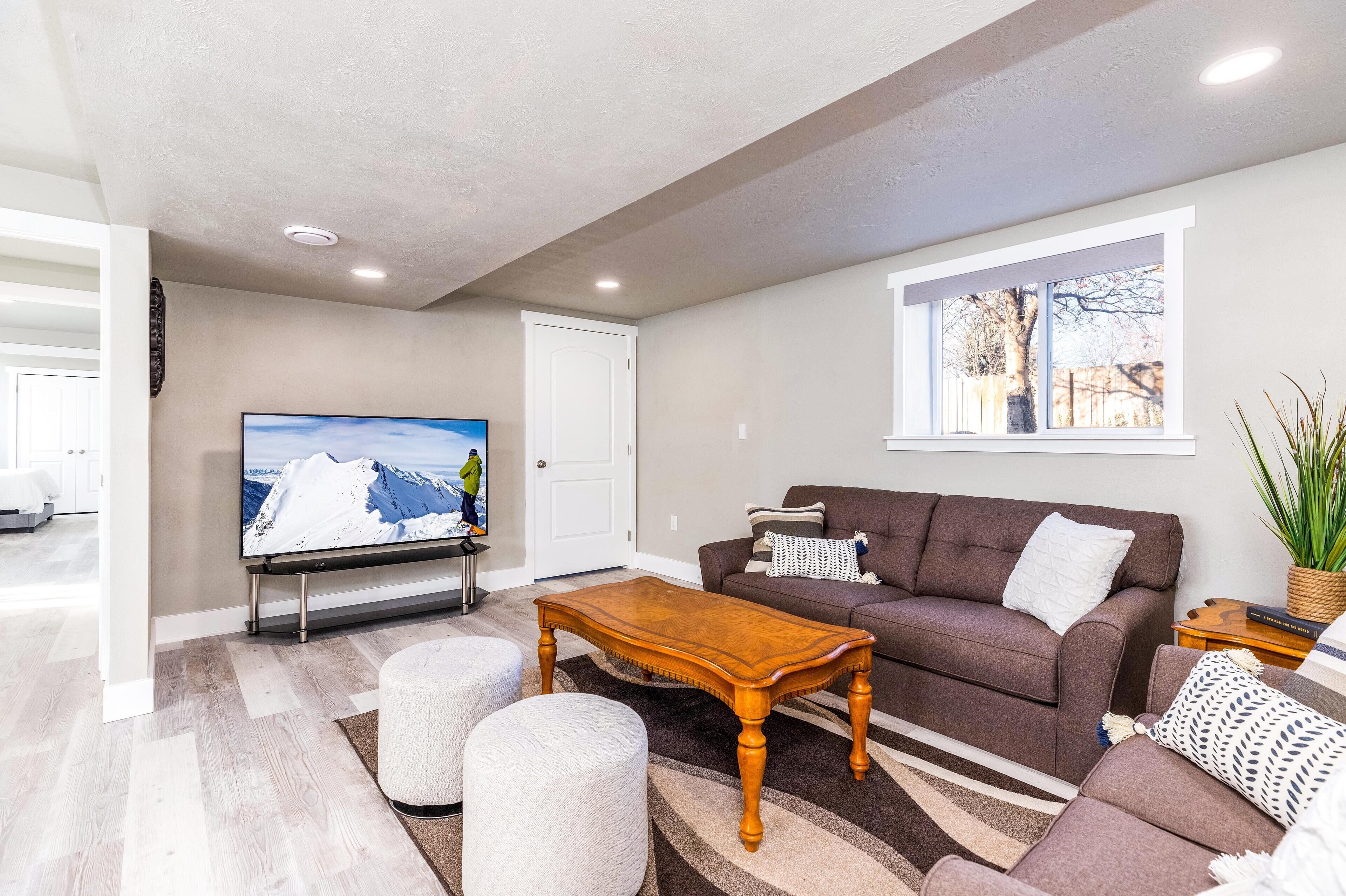 A modern living room with a flat-screen TV, a brown sofa, a wooden coffee table, and a large window with a view of trees.