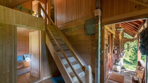 A wooden cabin interior with a staircase leading to a loft area, a door to a bedroom, and a view of the outdoors through a large window.