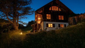 A rustic wooden house with a large staircase leading to the entrance, surrounded by tall trees and a grassy area.