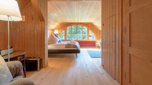 A cozy bedroom with a wooden ceiling, a large bed, a desk, and a window with a view of trees.