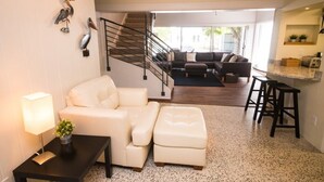 A modern living room with a sofa, ottoman, and a coffee table with a lamp. A staircase leads to a dining area with a bar and stools. - Lido Beach House
 (Sarasota)