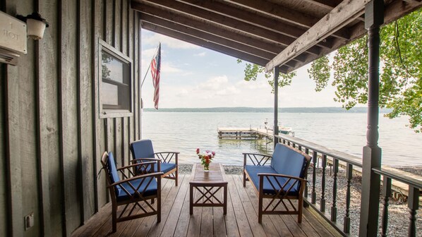 A wooden deck with two chairs and a table, overlooking a calm lake with a dock and a boat.