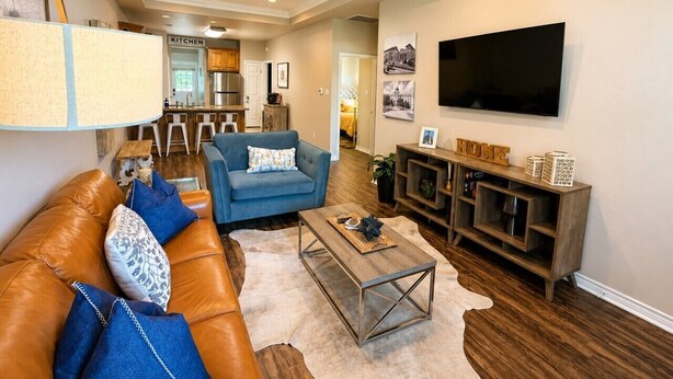 A living room with a sofa, armchair, coffee table, TV, and a kitchen area in the background.
