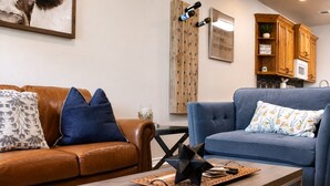 A living room with a brown leather sofa, a blue sofa, a wooden coffee table, and a kitchen area with wooden cabinets and a microwave.