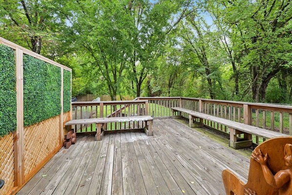 A wooden deck with benches and a wooden railing, surrounded by lush green trees.