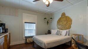 Slaapkamer met een bed, houten vloer, plafondventilator, raam met zonwering en een tafeltje met een plant.