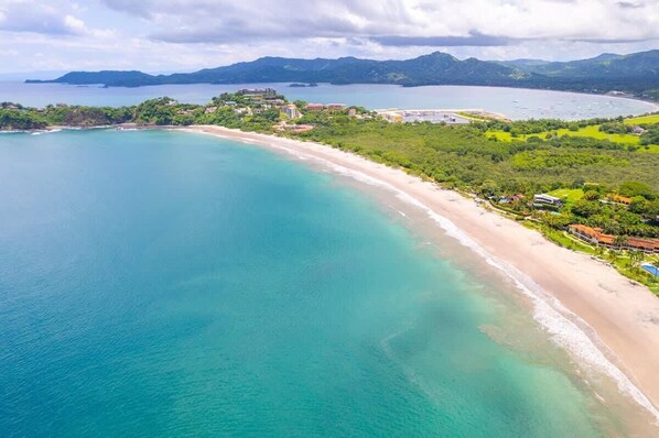Vista a érea de una zona costera con playa, vegetación exuberante y un gran complejo de edificios.