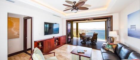 A spacious living area with a ceiling fan, a TV, a dining table with chairs, a sofa with cushions, and a large window offering a view.