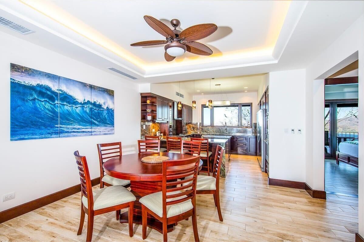 Un coin repas avec une table ronde et des chaises, un ventilateur de plafond et un grand tableau représentant une vague de l’océan au mur.