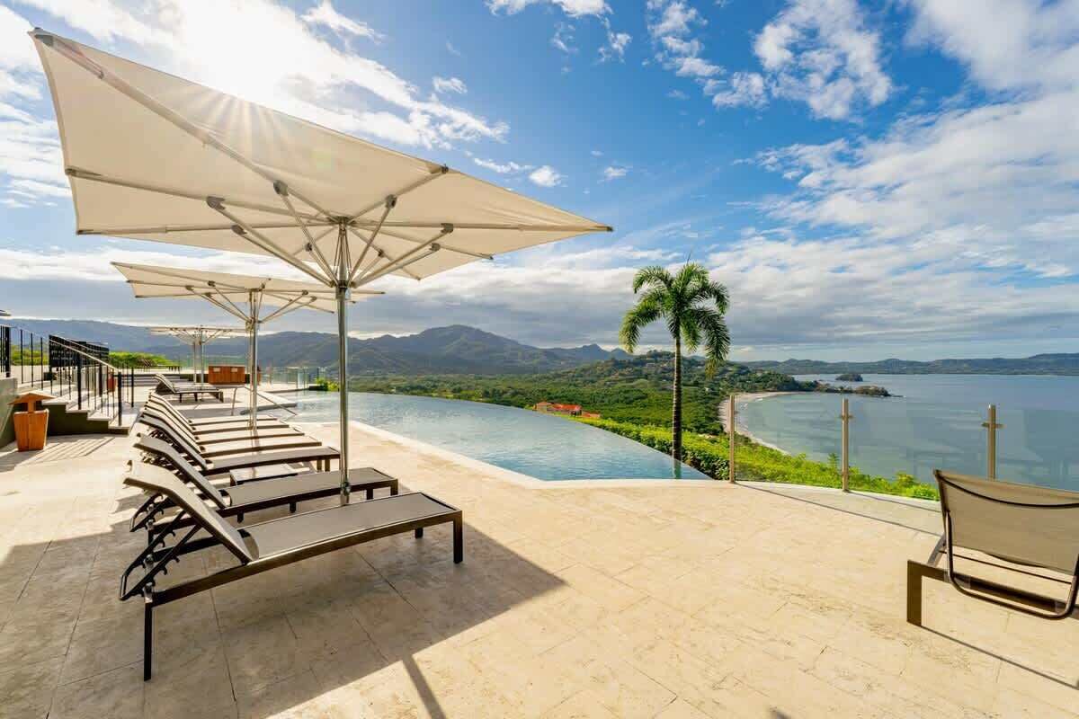 An infinity pool with a view of a body of water and mountains, surrounded by lounge chairs and umbrellas.