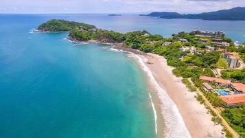 Eine Vogelperspektive einer Küstenlandschaft mit Strand, einer kleinen Insel und einem Resorthotel mit mehreren Gebäuden.