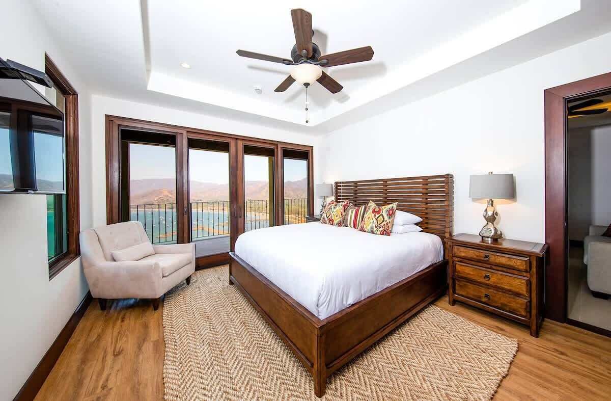 A bedroom with a large bed, a ceiling fan, a nightstand, a lamp, a chair, and a view of the mountains through the windows.