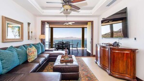 A modern living room with a sofa, ottoman, and a coffee table. There is a ceiling fan, a TV mounted on the wall, and a view of the outdoors through a glass door. - 360 Splendor 308F- 3 BR Condo, Breakfast Included (360 Splendor)